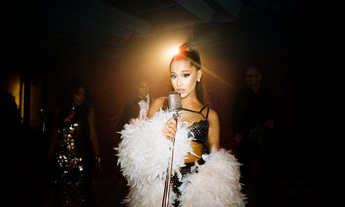 Glamorous recording artist, Ariana Grande, is holding a vintage microphone and wearing a black sequined top and boa in a studio.