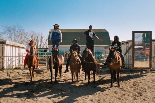 Outdoor shot of five people on horseback in sandy paddock, two standing, others riding, under a blue sky, with buildings and fencing visible.