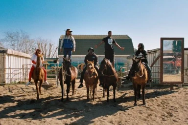 Five people on horseback in a sandy corral with a barn background, photographed by Pham.