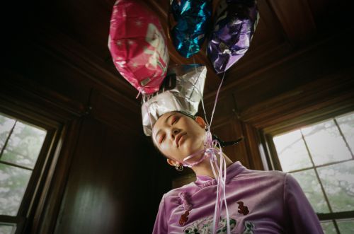 Low angle of Asian woman with closed eyes, wearing a purple dress, tied to colorful balloons in a dimly lit, paneled room.