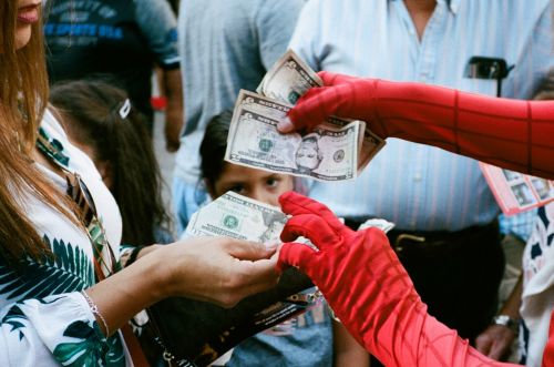 Person dressed as Spiderman receiving money from another person in a public outdoor setting with other people present in the background.