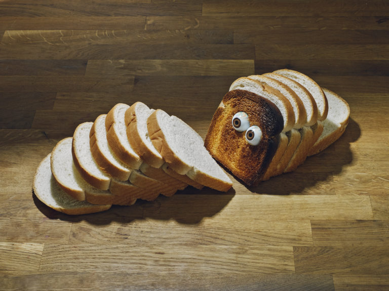 Slices of white bread, with a toasted slice featuring googly eyes, are arranged on a wood surface in a brightly lit, humorous still life.