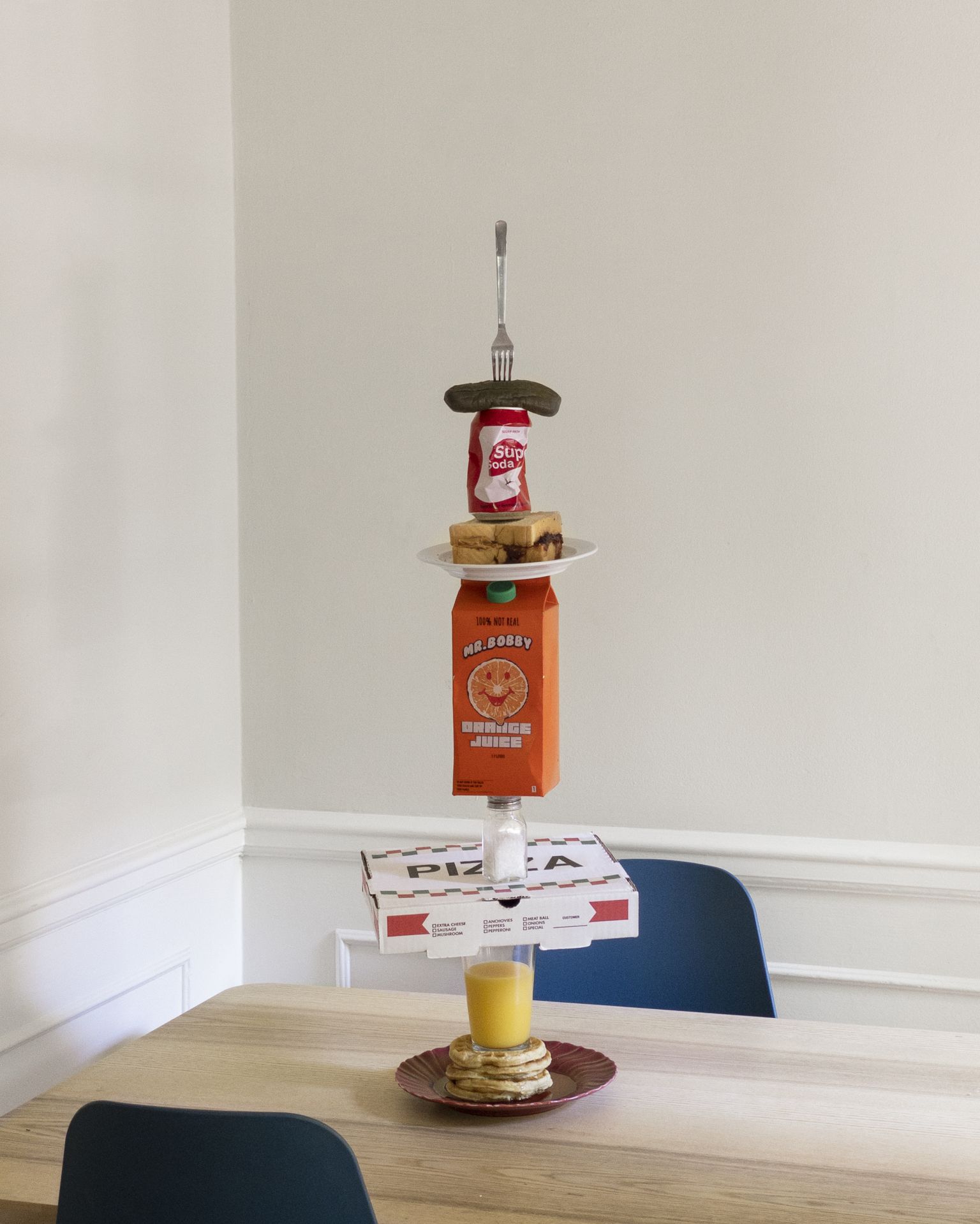 Stack of food and beverages on a wooden table indoors, featuring a pickle, soda can, sandwich, juice box, pizza box, and pancakes.