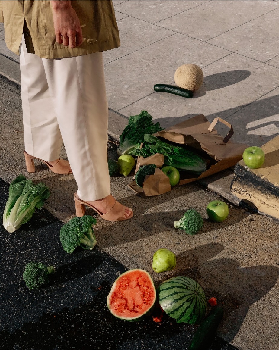 Woman in neutral tones stands near a collection of fruits and vegetables, including watermelon, apples, lettuce, and broccoli on a sidewalk.
