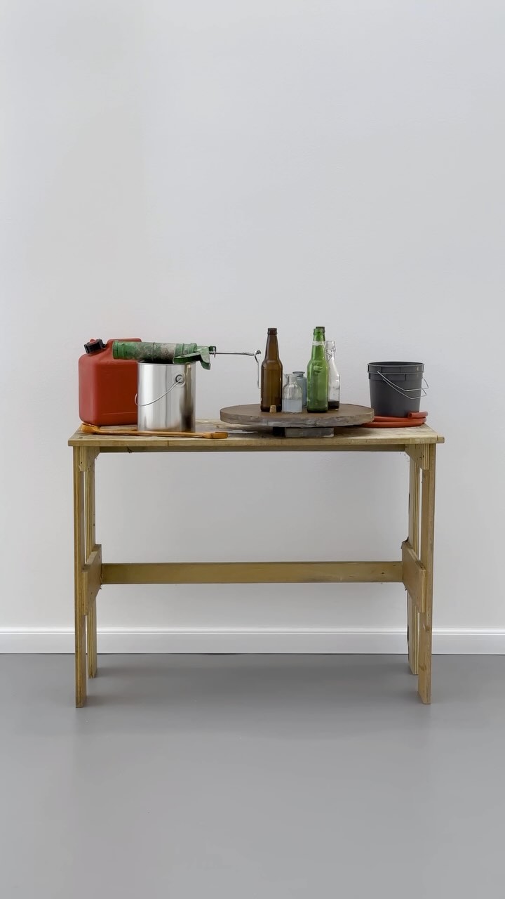 Still life shot of a wooden table holding bottles, a gas can, and paint supplies against a white wall with a grey floor.