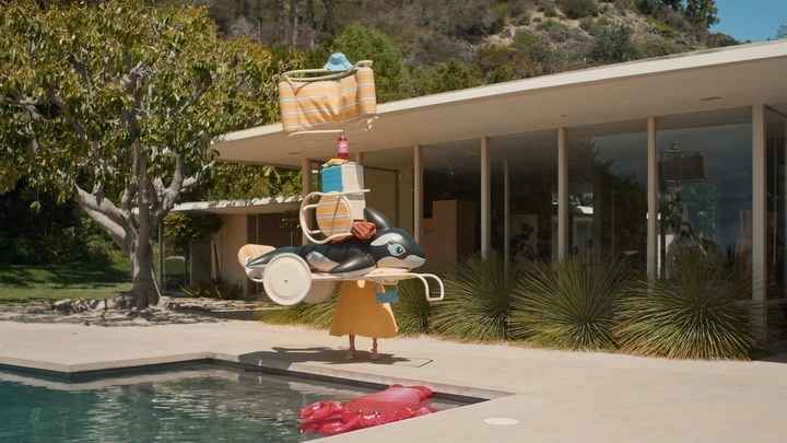 A playful scene outdoors: a person carries a whimsical tower of inflatable toys by a pool in front of a modern house against a leafy hillside.