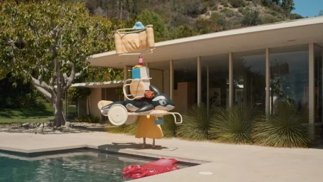 Person wearing a yellow dress balancing a towering stack of pool floats, chairs, and luggage on a rolling cart in front of a modern house.