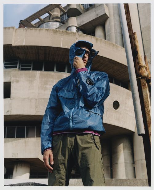 Adult male wearing a blue hooded jacket and sunglasses outdoors, standing in front of a tall brutalist concrete building on a sunny day.