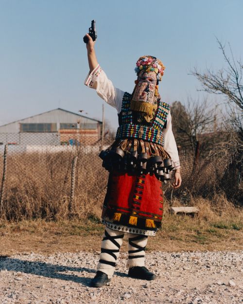 Person in ornate, colorful traditional clothing and mask, holding a pistol in the air on a gravel road with blurred background.