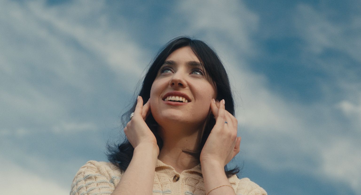 Smiling woman with dark hair touches face, looking up toward cloudy sky outdoors in medium shot with natural light in daylight.