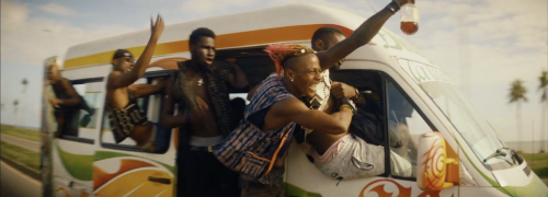 Excited group of diverse young adult friends cheer from the windows of a decorated van, traveling a sunny coastal road.
