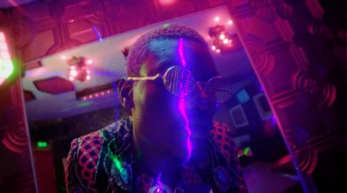 Portrait of a person wearing decorative glasses and patterned jacket, bathed in purple and green light in an indoor setting.