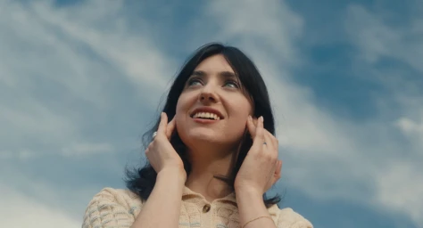 A smiling woman with dark hair looking upwards against a blue sky with soft white clouds, captured from a low angle.