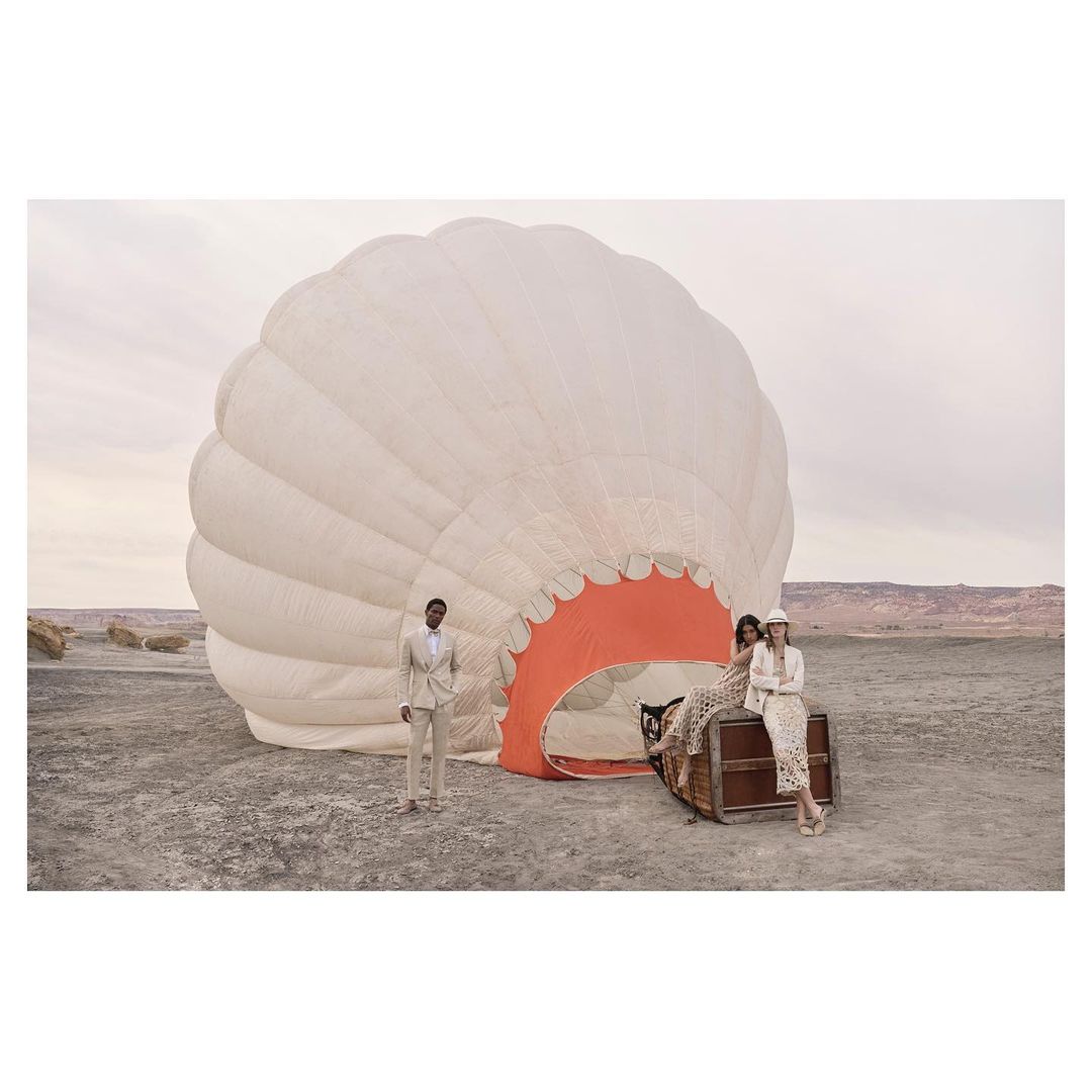 Fashion editorial image featuring three models in a desert landscape with a partially deflated hot air balloon and a vintage trunk.