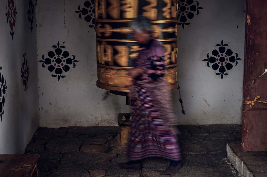 Blurred motion of elderly person spinning prayer wheel in a stone building with painted walls and dark stone floor.