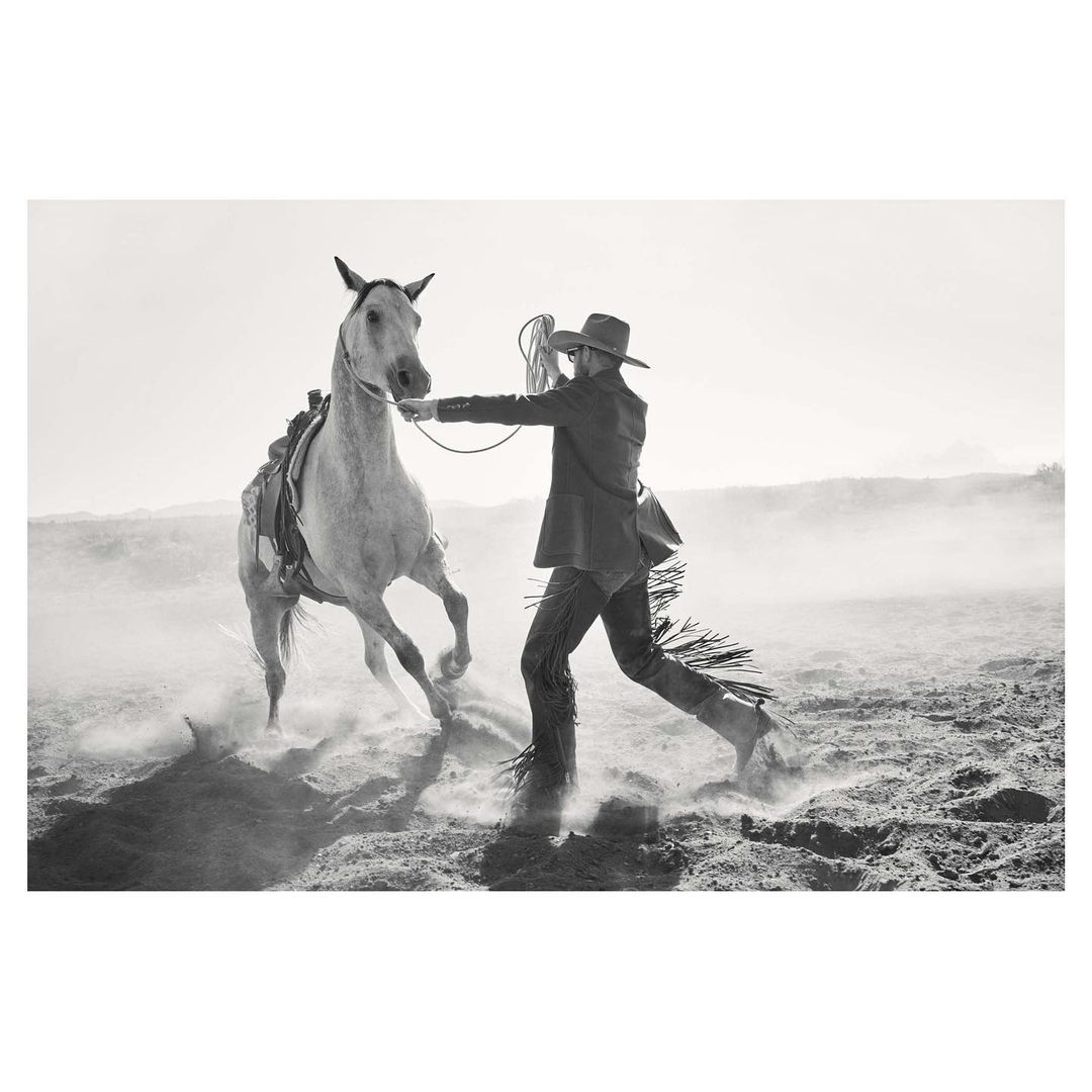 Monochrome image of a cowboy holding the reins of a rearing white horse in a dusty outdoor setting. The rider wears chaps, boots, and a hat.