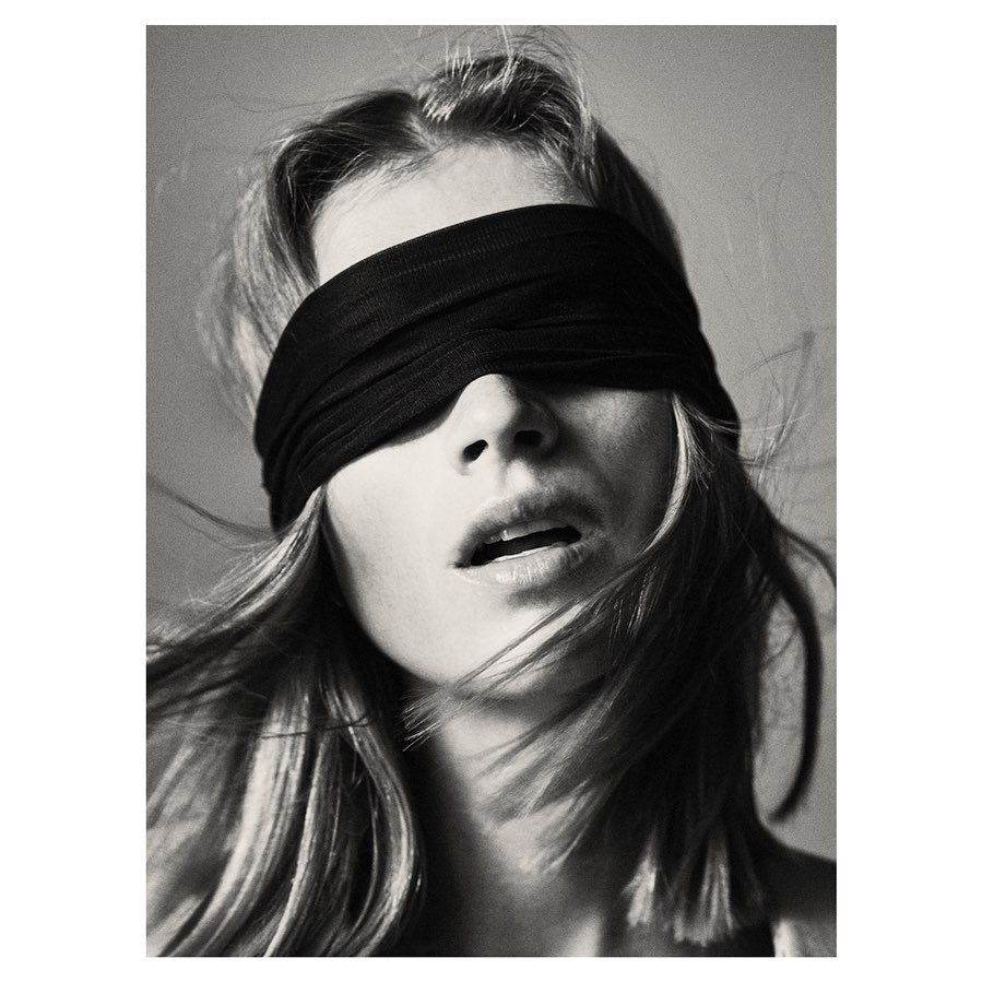 Monochrome portrait of a woman with a black blindfold, fair skin, and light hair. Studio shot with diffused lighting.