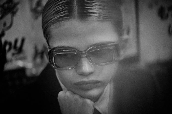 Close-up monochrome portrait of a model wearing thick-rimmed sunglasses and resting chin on hand by Alistair Taylor-Young.