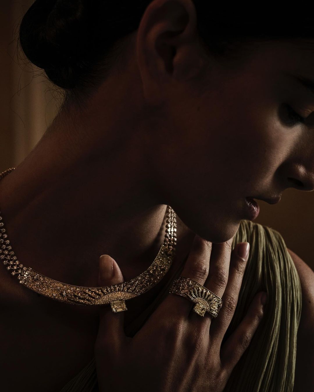 Woman showcasing a gold necklace and ring set with a subtle olive green dress, soft lighting, and a neutral backdrop.