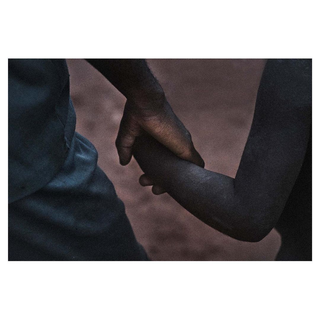 Close-up photograph features two individuals holding hands, depicting a connection and subtle intimacy in a simple composition.