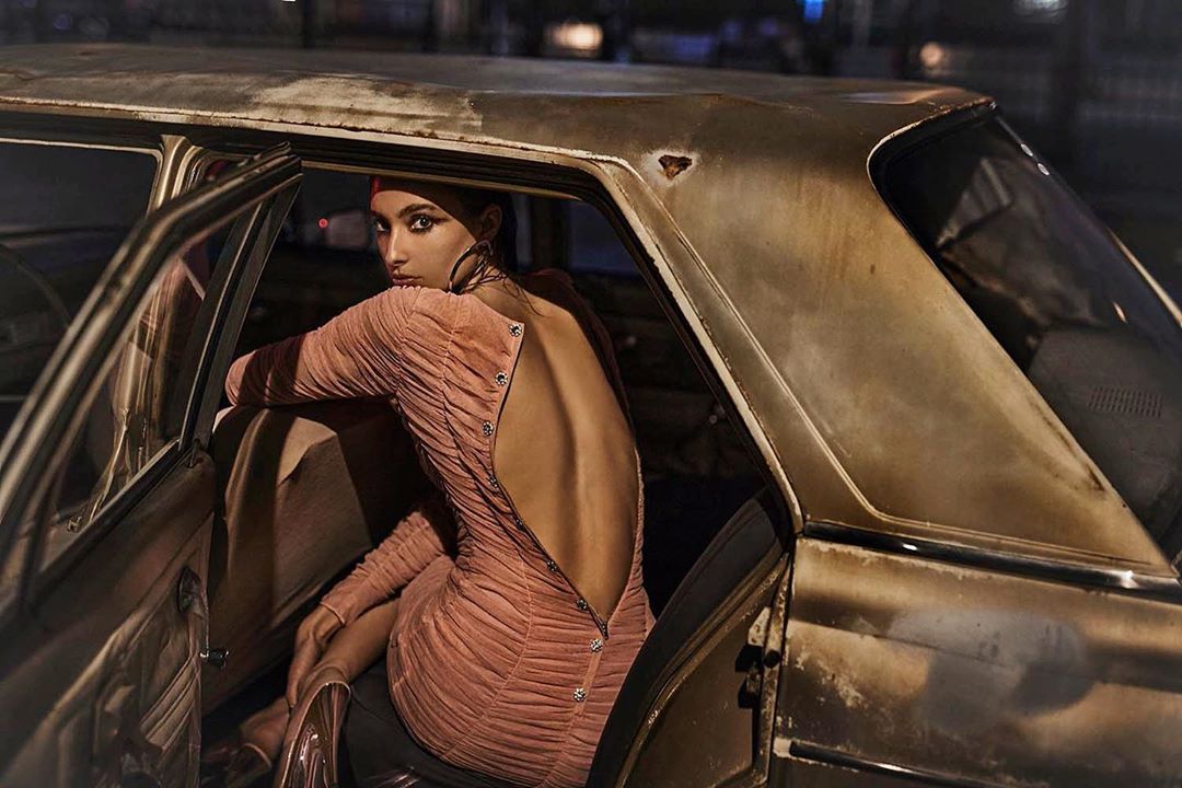 Glamorous woman in a pink dress sitting inside an old car with the door open, captured in an atmospheric nighttime outdoor fashion shoot.