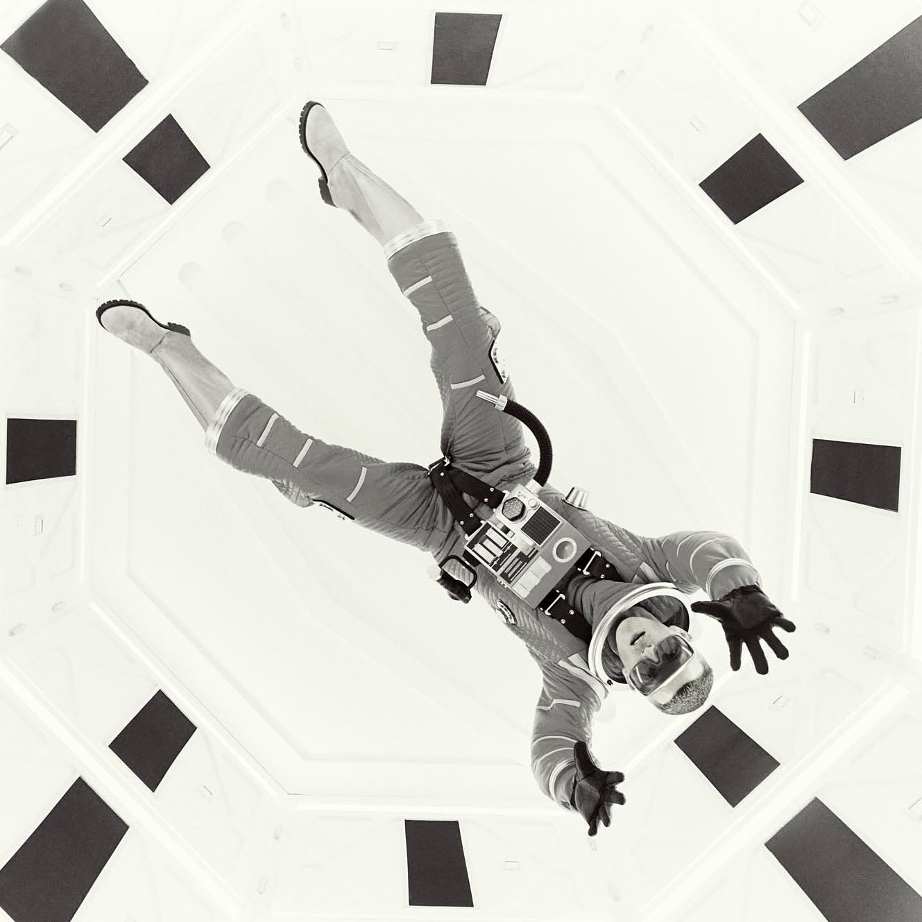 Monochrome shot of an astronaut in a space suit floating in a white, octagonal space station tunnel with geometric black accents.