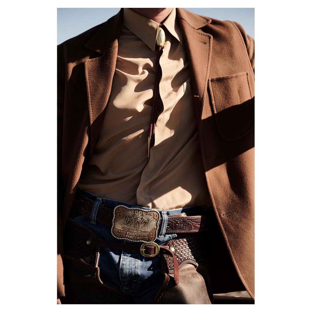 A close-up shot of a person wearing a brown jacket, beige shirt, blue jeans, and a large belt buckle with an outdoor background.
