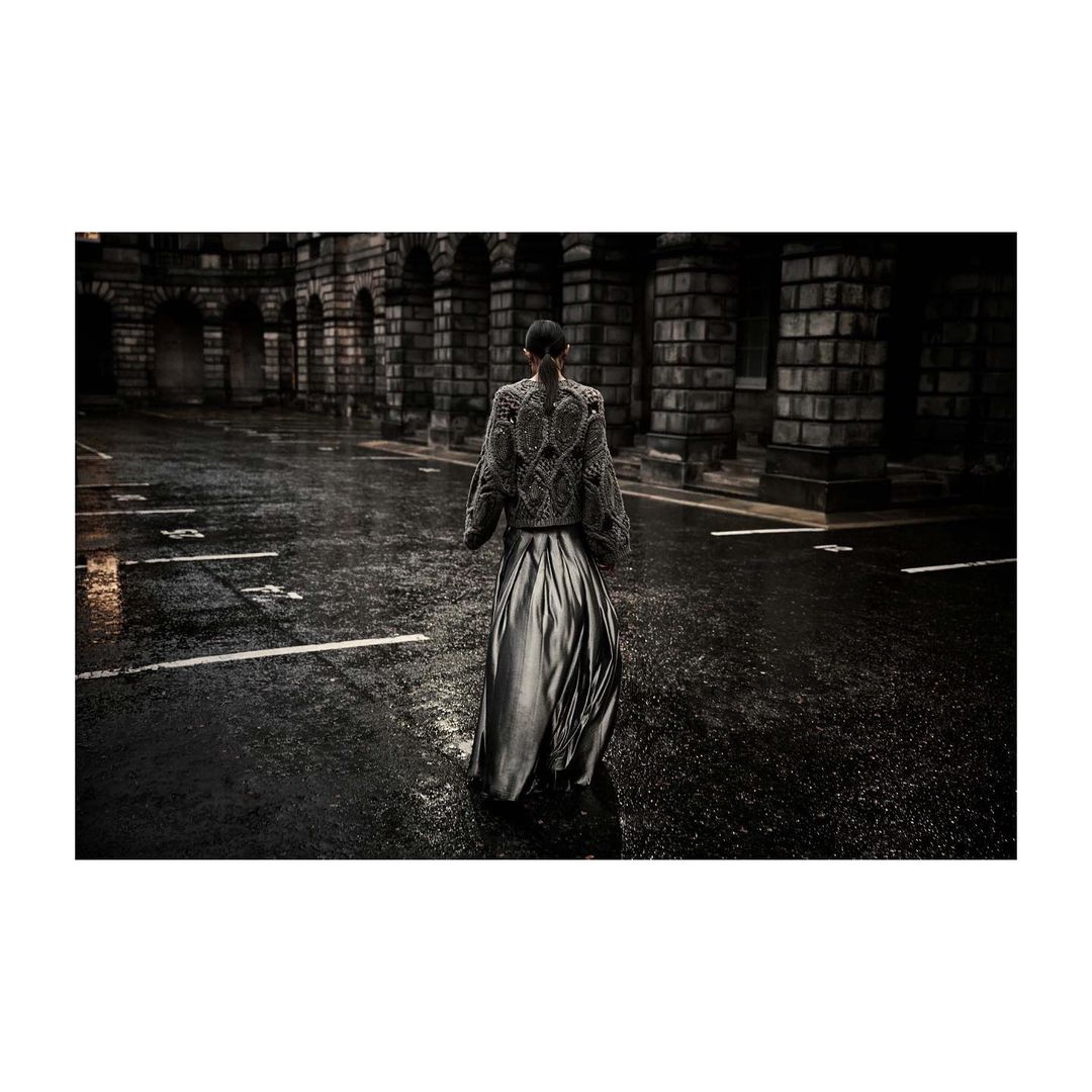 A person stands in a dark, wet street lined with stone buildings featuring arched entryways, wearing an embroidered jacket and long skirt.