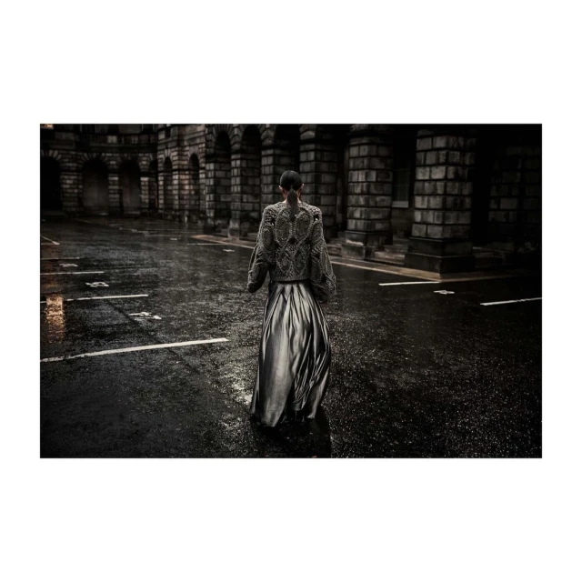 A person stands in a dark, wet street lined with stone buildings featuring arched entryways, wearing an embroidered jacket and long skirt.