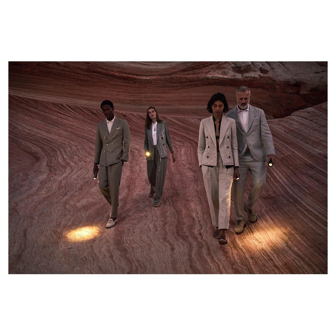 Four people in suits walk across a desert landscape carrying flashlights. The ground is patterned with bands of tan, beige, and brown.