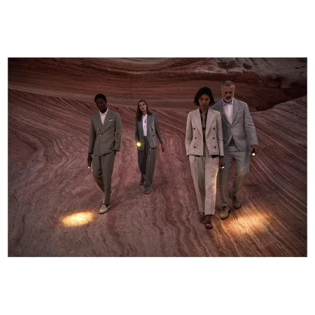 Four people in suits walk across a desert landscape carrying flashlights. The ground is patterned with bands of tan, beige, and brown.