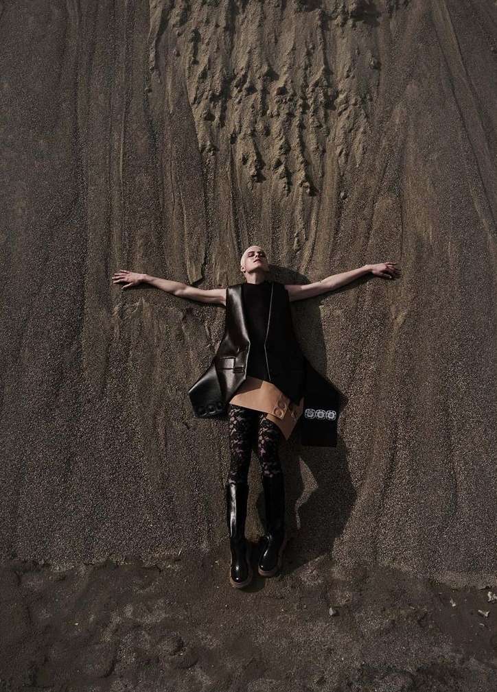 Model lying flat on a dark textured sand dune with arms outstretched, wearing dark tunic and boots.