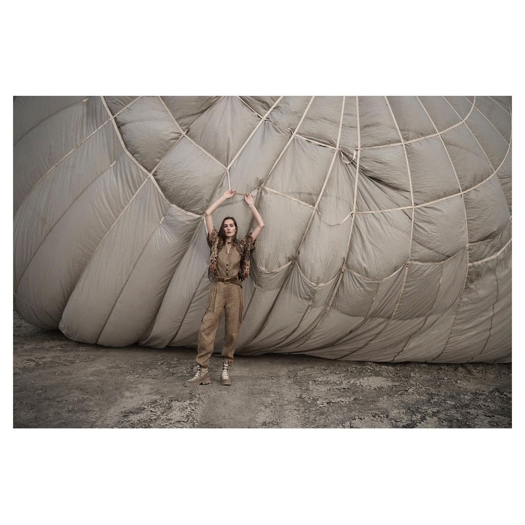 A woman in tan clothing poses in front of a large balloon-like structure on a grey surface, illuminated by daylight.