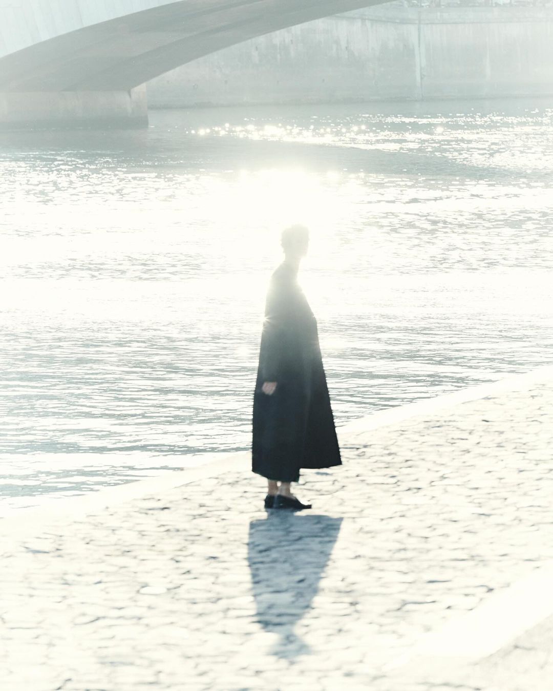 A person in a dark coat standing along a stone riverbank with bright sunlight reflecting off the water behind them.
