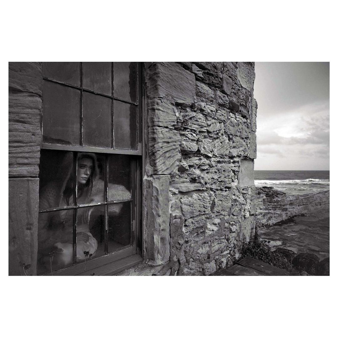 Monochromatic portrait of a woman looking out of a window in a stone building with a view of the ocean on an overcast day.
