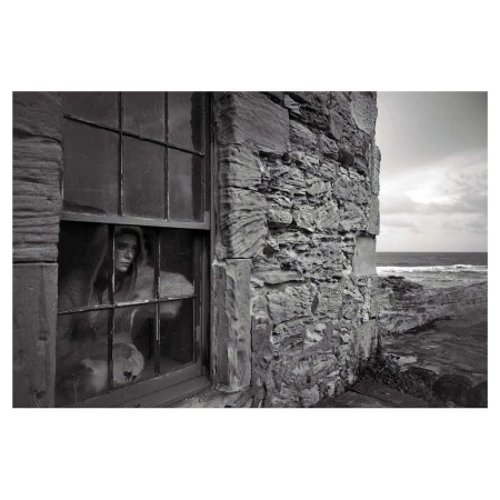 Monochromatic portrait of a woman looking out of a window in a stone building with a view of the ocean on an overcast day.