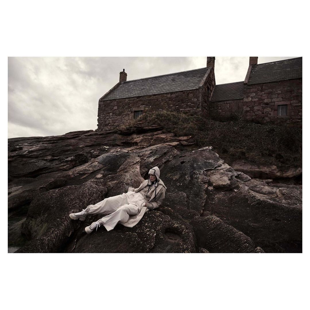 An adult in neutral clothing reclines on rocks by stone buildings under a cloudy sky, showcasing a contemplative fashion editorial outdoors.