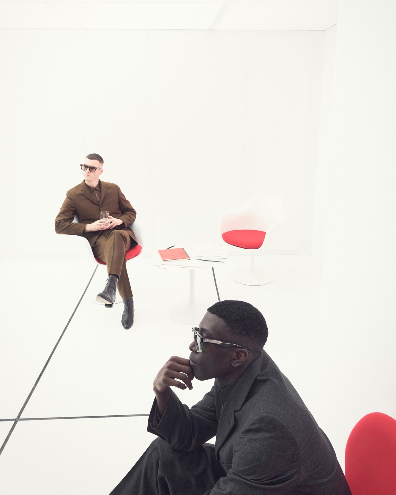Two stylish men in suits in a bright white room with modern red and white chairs, one holding a glass, in a high-contrast fashion editorial.