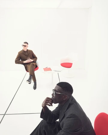 Two stylish men in suits in a bright white room with modern red and white chairs, one holding a glass, in a high-contrast fashion editorial.