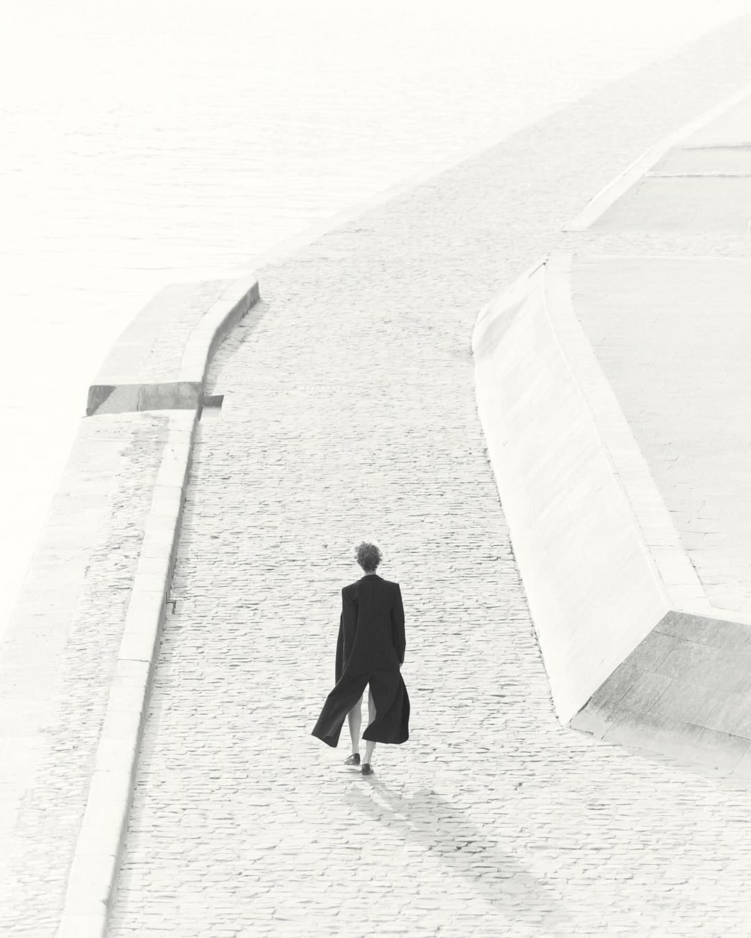 Back view of an adult in a dark coat walking on a paved surface near a retaining wall on a bright day; monochromatic color scheme.