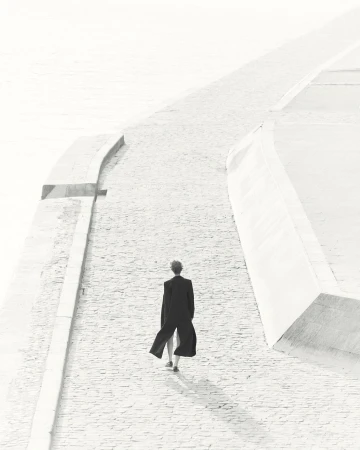 Back view of an adult in a dark coat walking on a paved surface near a retaining wall on a bright day; monochromatic color scheme.