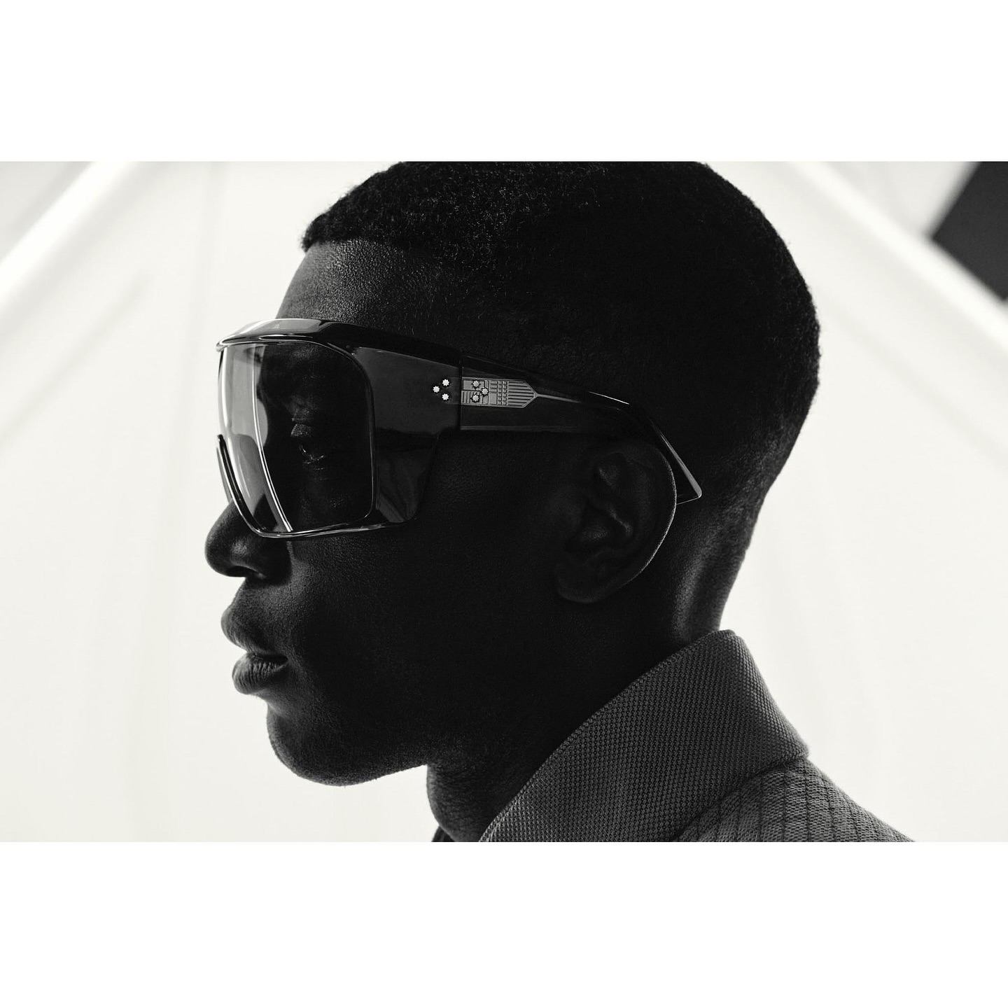 Profile portrait of a dark-skinned man wearing futuristic black visor sunglasses and a grey collared shirt against a light background.
