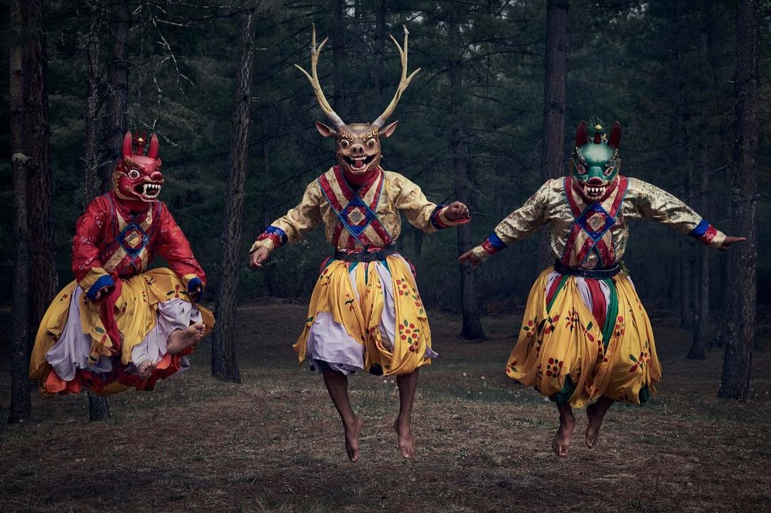 Three figures in ornate costumes and animal masks leap in a forest, creating a surreal and captivating visual narrative.