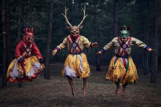 Three figures in ornate costumes and animal masks leap in a forest, creating a surreal and captivating visual narrative.