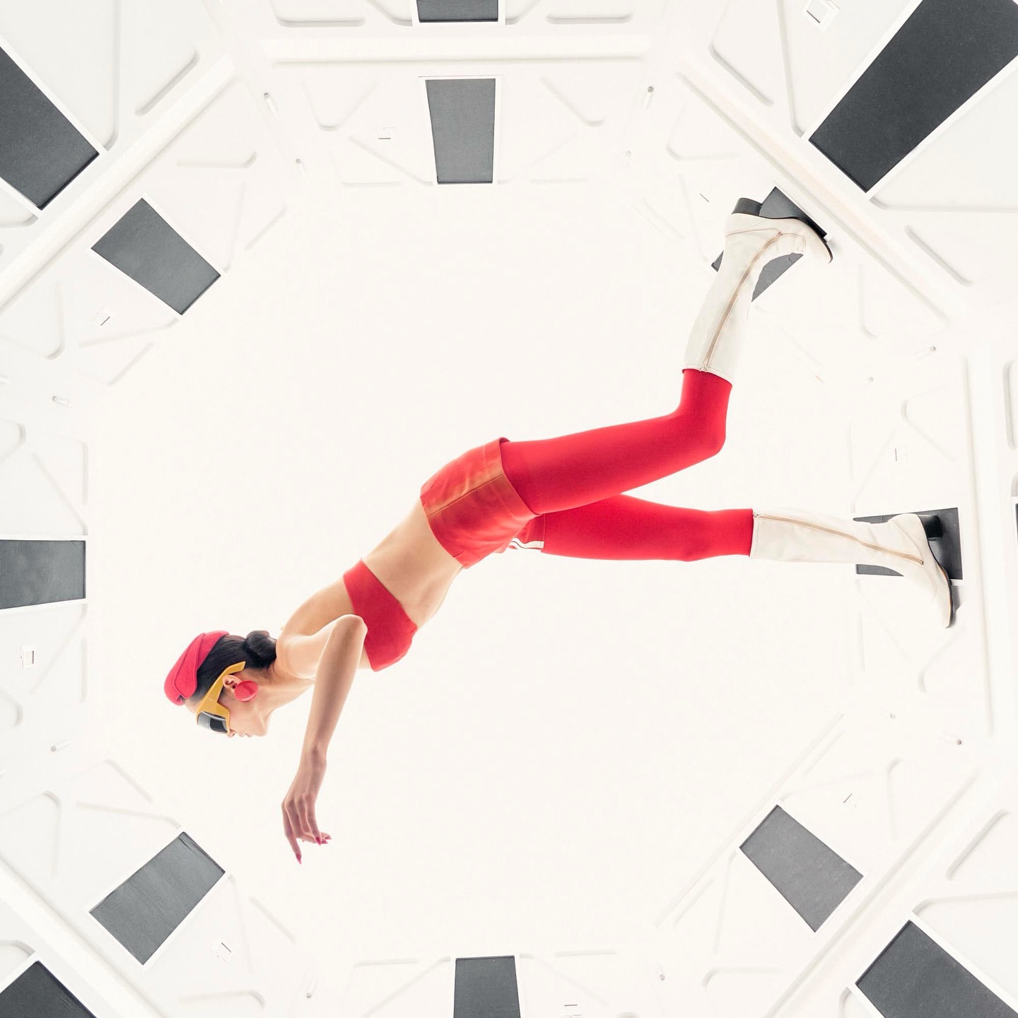 Woman in red top, shorts, leggings, and white boots poses in an octagon-shaped, light-filled structure with gray and white panels.