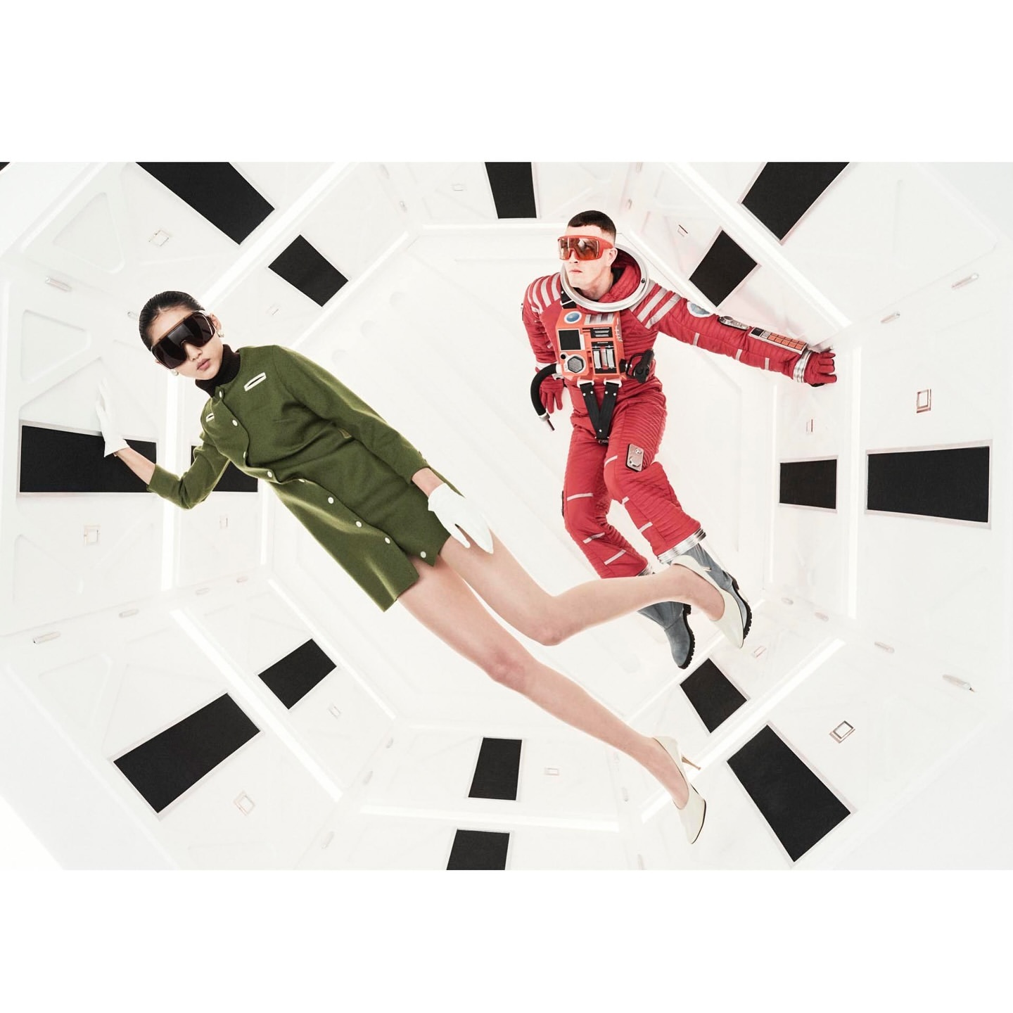 Two people in futuristic costumes pose in a brightly lit, white geometric interior space with black accents. 