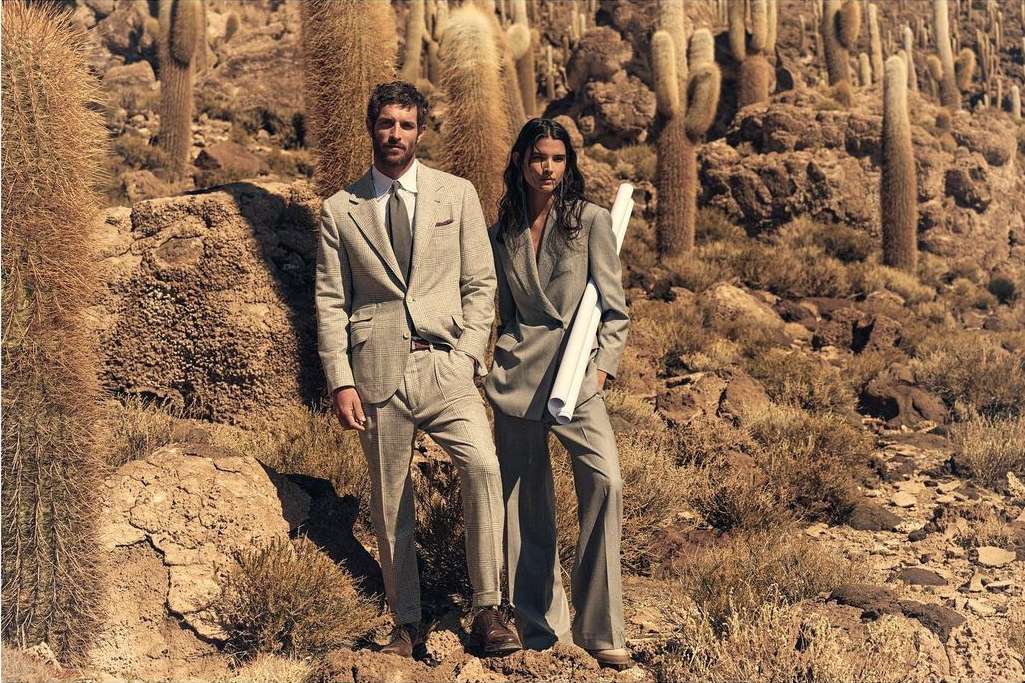 A stylishly dressed couple stands in a desert landscape with tall cacti and rocky terrain, wearing grey suits in an outdoor fashion shot.