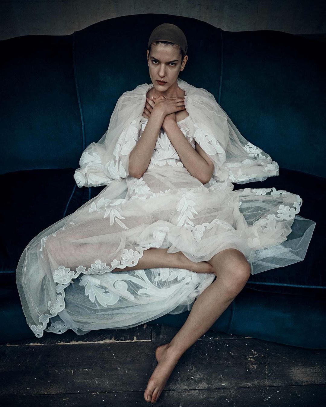 Woman in an ornate white dress and cap, seated on a blue velvet couch, hands crossed, looking at the camera in a studio setting.
