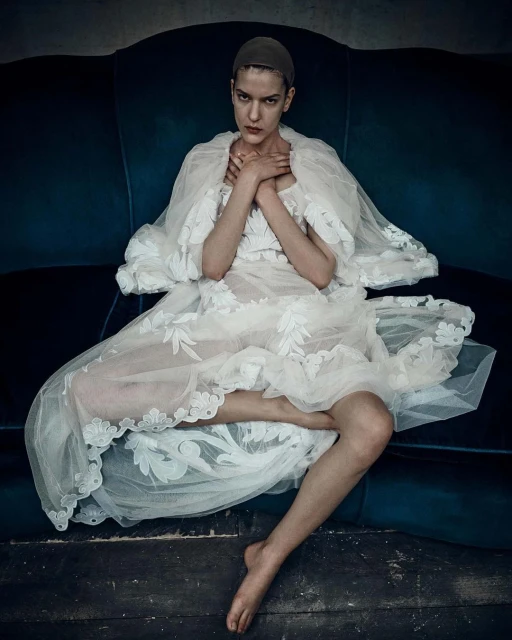 Woman in an ornate white dress and cap, seated on a blue velvet couch, hands crossed, looking at the camera in a studio setting.