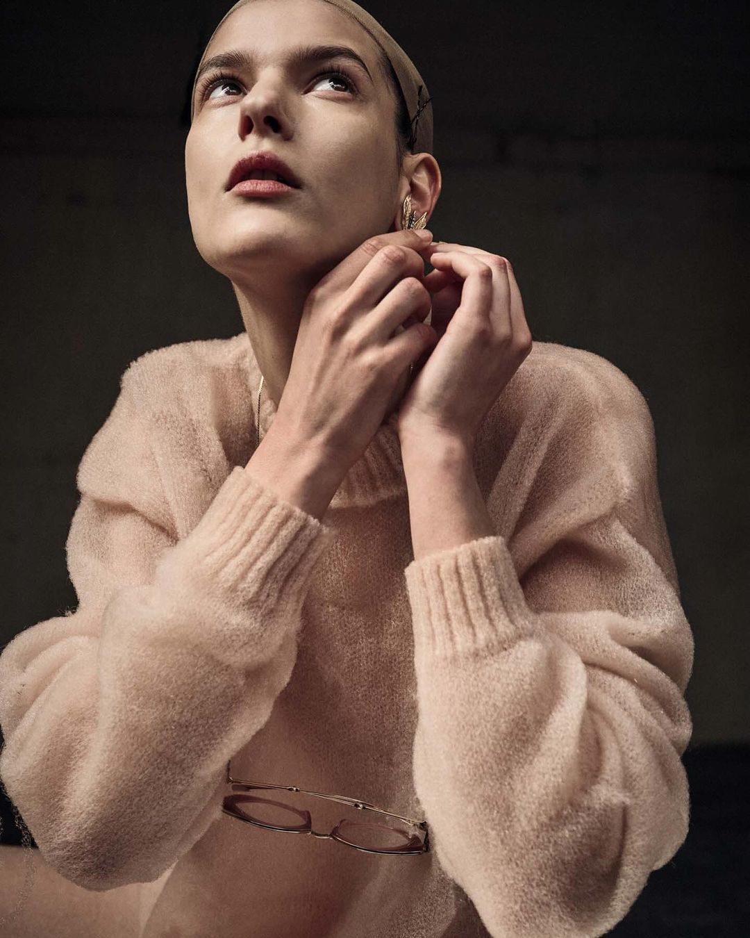 Woman in beige turtleneck sweater adjusts earring, gazing upward in dimly lit setting. Focus on fashion and quiet mood.